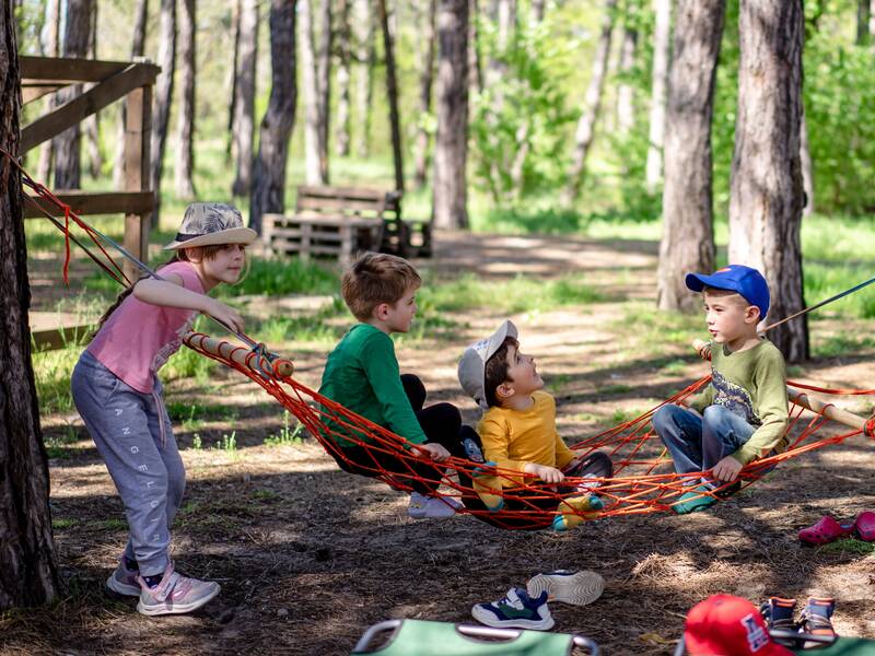 Drei kleine Jungs sitzen auf einer Netz-Hängematte im Wald zwischen vielen Bäumen. Ein Mädchen steht links daneben und hält sich an der Hängematte. 