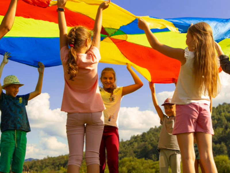 Mehrere Kinder stehen in einem kreis und halten ihre Hände nach oben. Sie halten alle zusammen ein großes, rundes, buntes Tuch über ihre Köpfe. Sie schauen sich dabei unter dem Tuch durch an. 