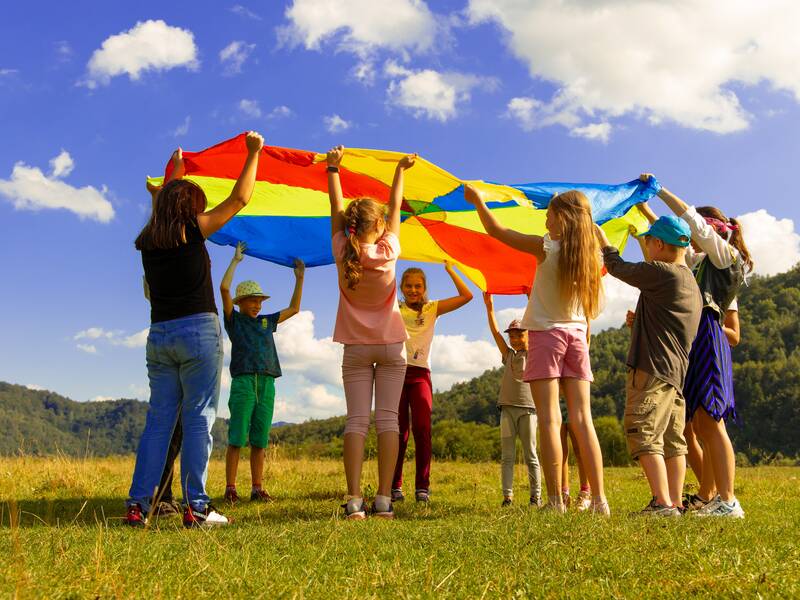 Kinder die im Kreis stehen und nach oben ein großes, buntes und rundes Tuch halten. Sie schauen sich unter dem Tuch durch an. 